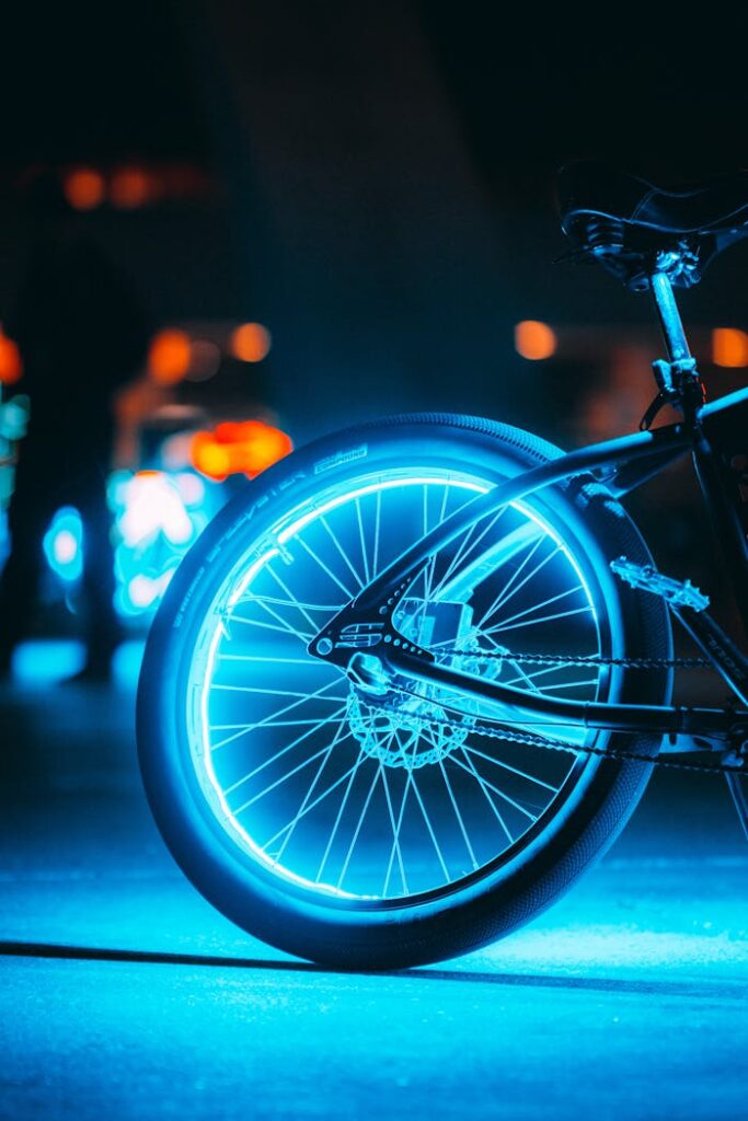 A close-up of a glowing bicycle wheel on a city street at night, showcasing neon lights.